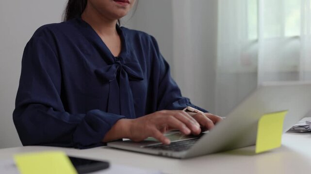 Female office employee typing rapid text on silver laptop computer keyboard while sitting beside stacked corporate documents and files at white desk in bright workplace setting