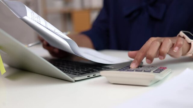 Close view of female hands operating laptop trackpad and pressing keys while reviewing corporate invoice sheets with calculator on white office work table during business day
