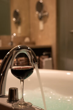 Glass of red wine by a filling bathtub with foam.