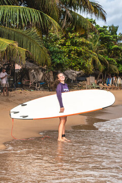 Young smiling woman standing on a tropical beach holding a white surfboard under palm trees