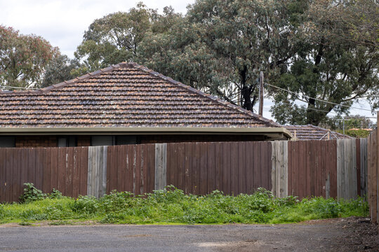 Weathered wooden fence with patch repairs surrounding an older home, featuring unkempt grass along the property boundary. Low income neighborhood in Australia.