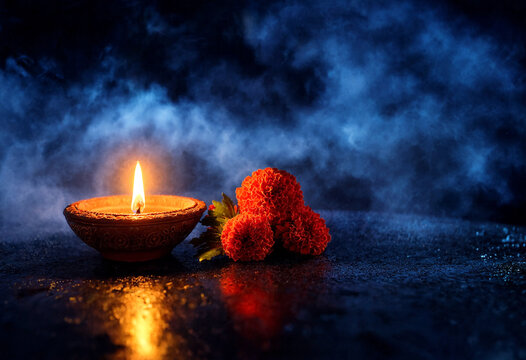 Traditional clay diya lamp with burning flame and red flowers on dark moody background