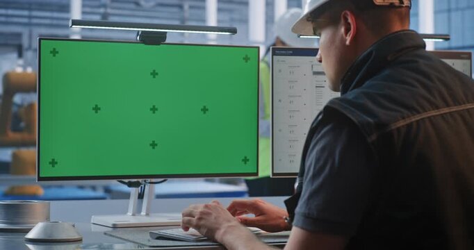 Industrial Worker in Safety Gear Sits at Control Desk Typing, Facing Glowing Green Screen Monitors. Displays Feature Trackers For Clean Chroma Key Compositing in Smart Factory. AI Manufacturing.