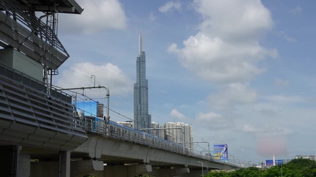 Elevated metro train passing station with Landmark 81 skyscraper in the background under a cloudy blue sky, Ho Chi Minh City, Vietnam
