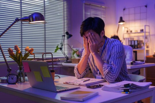 Exhausted Asian businessman covering face at office desk late at night for workplace stress and burnout concept