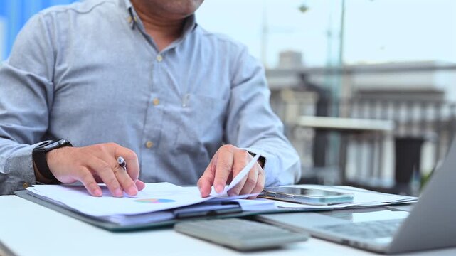 Businessman performs financial analysis reviewing corporate report, conducting market analysis over paper charts, completing data analysis near office report.