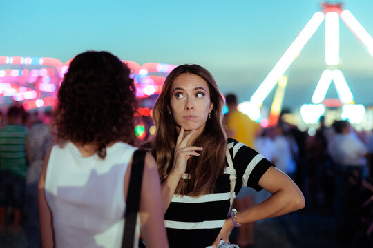 Puzzled Pensive Woman Looking At Friend At Night Amusement Park. Confused thoughtful female stares blankly towards her companion inside blurry festive fairground