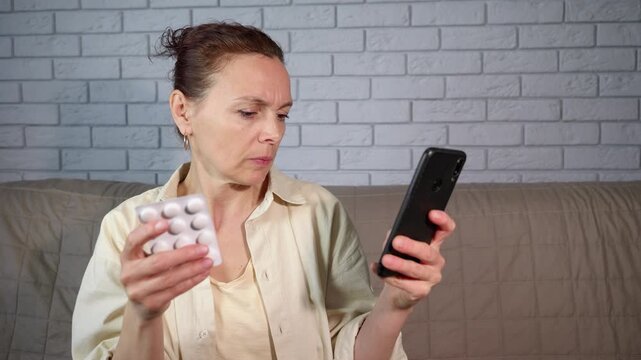 Mature woman buying pills online from home. Concerned middle-aged woman sitting on the sofa, holding a blister pack of pills and looking up medical information on her smartphone
