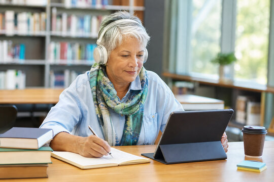 Older Woman Learning Independently with a Tablet and Headphones in a Library, Taking Digital Notes, Exploring Online Education, and Building Confident Technology Skills