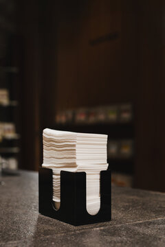 Stack of white tissue paper napkins inside a sleek black holder placed on a stone table in a cafe.