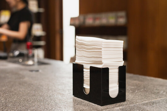 Stack of white tissue paper napkins inside a sleek black holder placed on a stone table in a cafe.