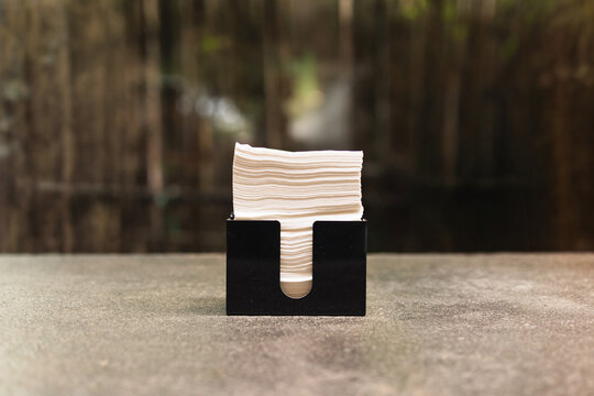 Stack of white tissue paper napkins inside a sleek black holder placed on a stone table in a cafe.