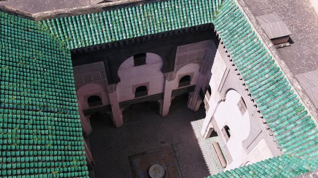 Elevated high-angle drone perspective looking down into the ornate inner courtyard, arches, and student cells of a historic madrasa framed by green glazed tile roofs in Fez, Morocco.