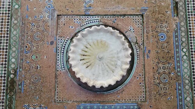 Direct vertical drone perspective capturing the fluted marble basin, ancient zellige mosaic tiles, and historic stone floor of a madrasa courtyard in Fez, Morocco.