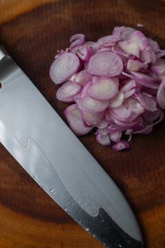 Vertical close up of chef knife blade beside sliced purple shallots on wooden board.