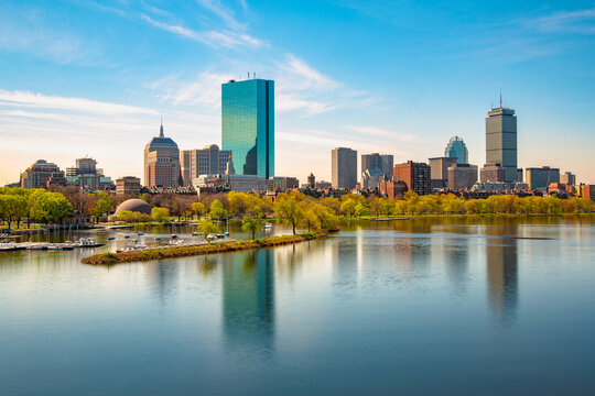 A vibrant daytime view of the Boston skyline featuring John Hancock Tower and Prudential Center, with reflections in calm waters of the Charles River.