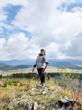 Senior woman hiker with red trekking poles on rocky hilltop. Smiling at mountain valley with pine forest under cloudy sky. Active aging, Nordic walking and outdoor tourism concept.