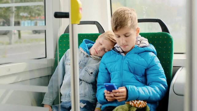 Two young boys seated side-by-side on green patterned bus seats, older boy wearing bright blue puffer jacket and striped hoodie focused on smartphone held with both hands while younger boy in light