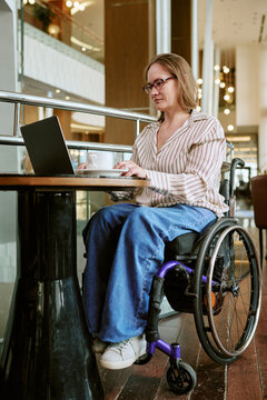 Vertical shot of middle aged Caucasian woman with disability sitting in wheelchair working on laptop in hotel lobby. Image suitable for remote work and digital accessibility advertising