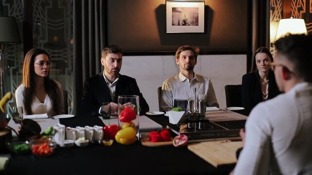 Seated panel of four adults faces presenter across long dining table in upscale private dining room while subtly shifting gaze and posture, conveying attentive evaluative mood, with meeting, dining