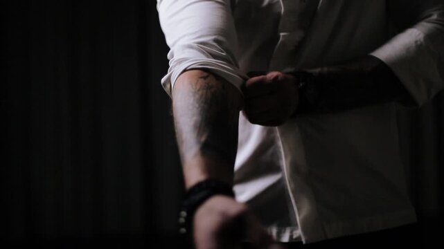 Close-up motion shot of torso and arms as person uses right hand to roll up white long-sleeve shirt, exposing tattooed left forearm, visible action includes cuff being gripped and pulled upward with