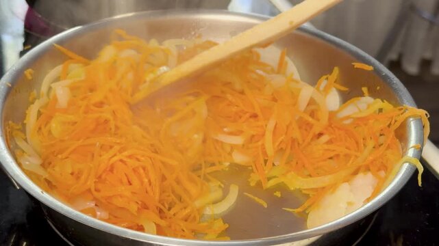 Sliced onion and grated carrot stirred while frying in pan on kitchen stove.