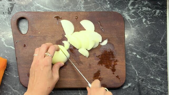 Hands slicing fresh white onion on wooden cutting board, overhead food preparation.