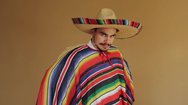 Medium close three-quarter portrait of young man wearing wide-brimmed braided straw sombrero with multicolor woven trim and bright horizontal striped serape poncho, posed against warm tan seamless