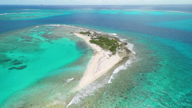Aerial drone footage shows waves breaking on both sides of tiny sandy Petit Tabac island encircled by extensive coral reef and turquoise sea