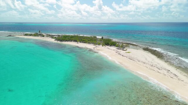 Drone pans slowly around narrow sandy Petit Tabac island stretching into open sea, framed by clear turquoise water and surrounding coral reef