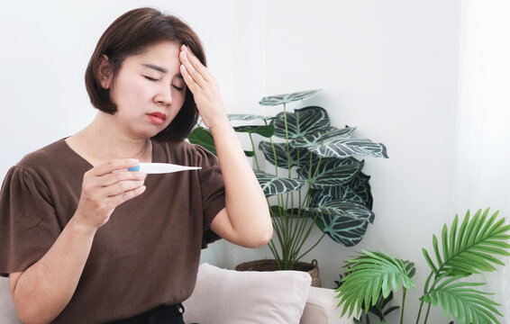 Sick Asian woman holding a digital thermometer touching her forehead with high fever, headache, flu or virus illness at home. Portrait of female patient feeling unwell in healthcare 
