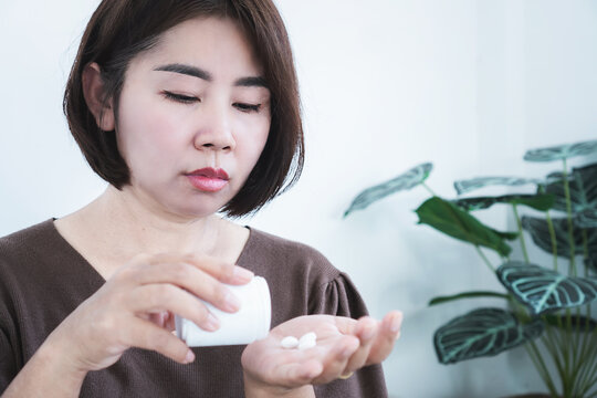 Asian woman pouring pills from bottle into hand for medical treatment, medication dosage, or supplements at home. Close-up portrait of female patient taking prescription 