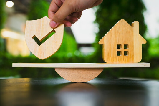Home insurance and property protection balance concept with hand placing wooden shield on seesaw scale opposite house model against green bokeh background for real estate risk equilibrium.