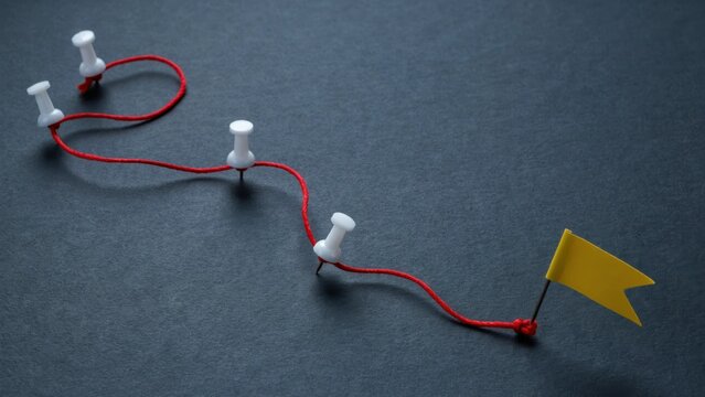 A red thread connected by pins leads to a yellow flag marker on a dark textured background. Metaphor for project planning, logistics, or reaching goals.