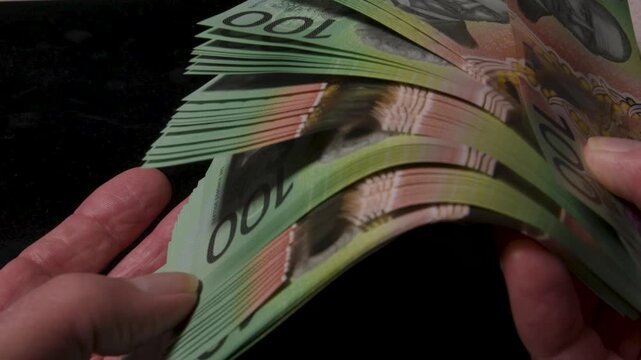 Close shot of human fingers riffling a thick stack of Australian 100 notes on a black background for money and finance themes.