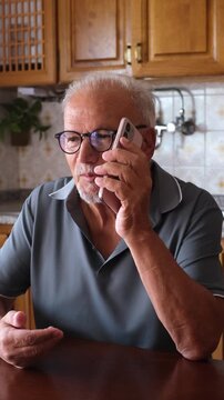Concerned elderly man with eyeglasses speaking on his mobile phone at home, gesturing with his hand during an intense and serious conversation while sitting at his kitchen table