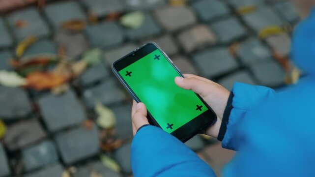 Close over--shoulder shot of child wearing bright blue padded jacket pressing thumb on smartphone with green chroma screen marked by four black tracking crosses, set against blurred cobblestone
