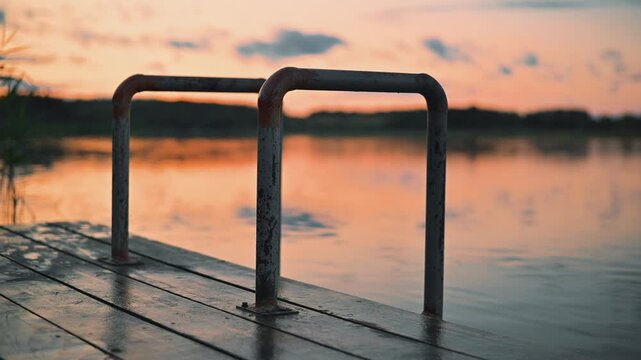 metal swimming pool ladder placed on a wooden deck by the water's edge. The scene captures a sunset with vibrant colors reflecting on the still water
