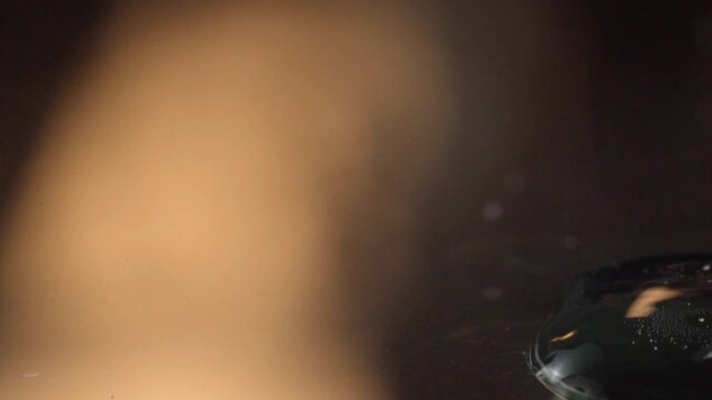 Clear liquid is dripping onto a glossy black surface in macro closeup. Smooth reflections and warm highlights emphasize the flowing liquid and polished surface texture.