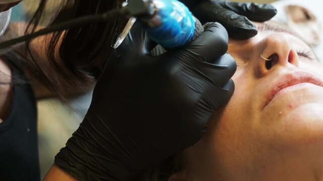 PMU artist in black nitrile gloves applies pigment along a woman's eyebrow using a precision tattoo machine during permanent makeup, static extreme close-up.