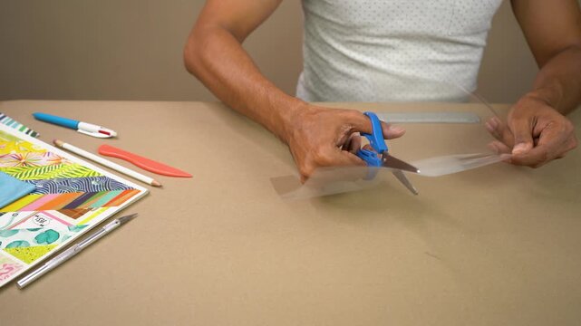 Person cutting clear acetate sheet with scissors at craft table indoors