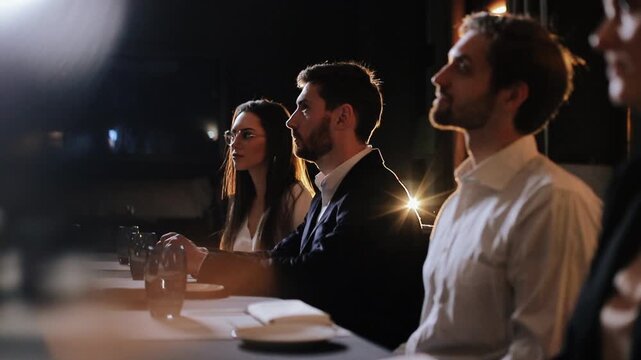 Three young professionals seated in row shown in right-side profile at dim indoor table, center subject bearded man in dark jacket and white shirt and left subject woman with long hair and glasses