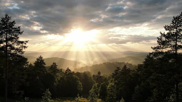 Sunbeams breaking through clouds over a misty forest landscape at sunrise.