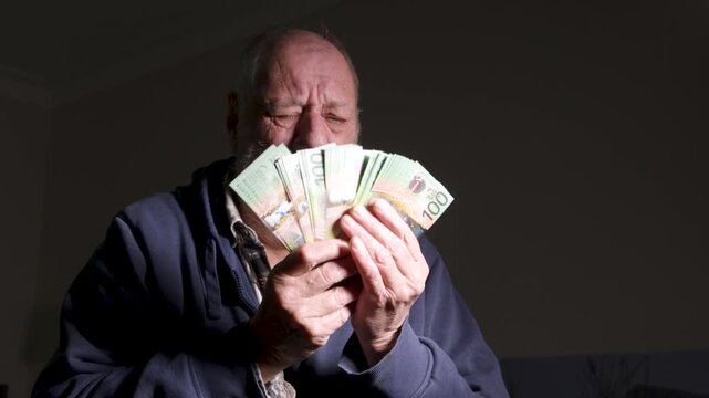 Bearded senior man shifts from joy to tears while clutching a fan of paper money under harsh indoor lighting.
