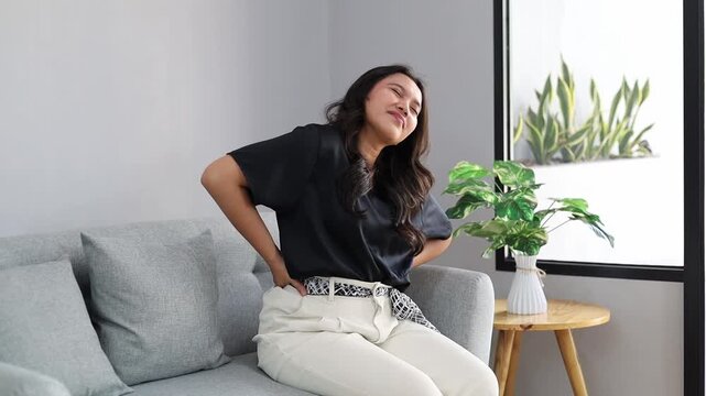 Pained Young Asian female experiencing lower back pain and discomfort while sitting on the couch at home