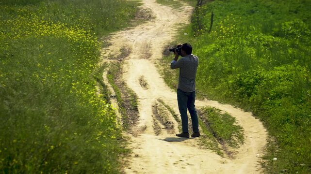 A photographer stands on a winding rural path beside a mustard field
