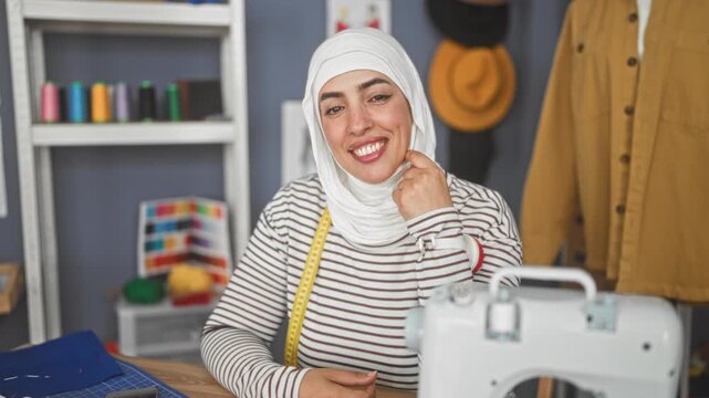 Woman smiling and touches hijab with hand, measuring tape around neck and sewing machine on table in studio; confident creative entrepreneur.