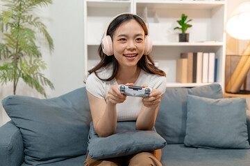 Young woman wearing wireless headphones while playing video games with a game controller at home. Excited gamer enjoying online gaming, entertainment, competition and modern digital lifestyle. © Imagestoyou
