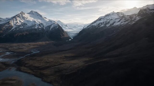 Cinematic aerial landscape of mountain glacier valley with winding glacial river, snow covered peaks and dramatic cloudy sky creating remote natural background of quiet grandeur and awe