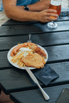 Plate of hearty food with sausages, French fries, creamy sauce, a breaded fried cutlet, and a roasted meat portion on a black outdoor table beside a glass of amber beer